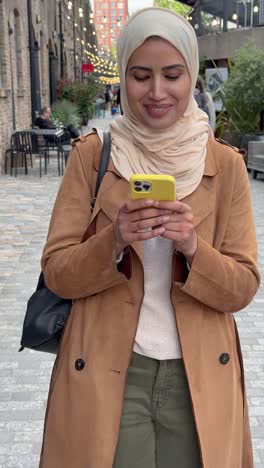 Vertical-Video-Of-Muslim-Woman-Wearing-Hijab-Walking-Along-City-Street-Sending-Text-Message-To-Date-On-Mobile-Phone